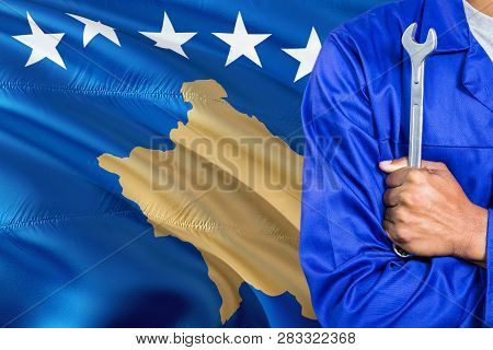 Mechanic In Blue Uniform Is Holding Wrench Against Waving Kosovo Flag Background. Crossed Arms Techn