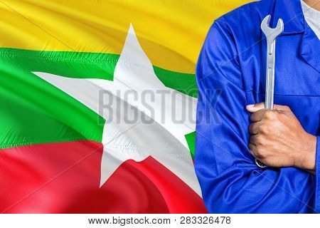 Burmese Mechanic In Blue Uniform Is Holding Wrench Against Waving Burma Flag Background. Crossed Arm