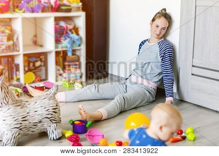 Tired Of Everyday Household Mother Sitting On Floor With Hands On Face. Kid Playing In Messy Room. S