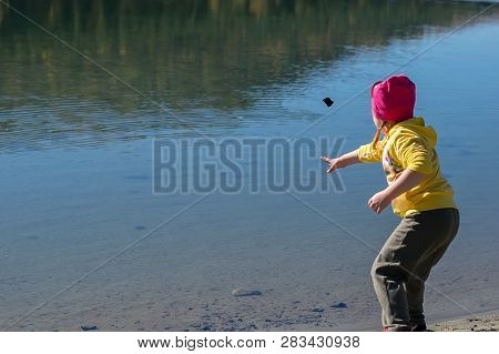 A Little Girl, A Child, In Warm Clothes And A Red Cap Throws Stones Into The Water From The Sandy Sh