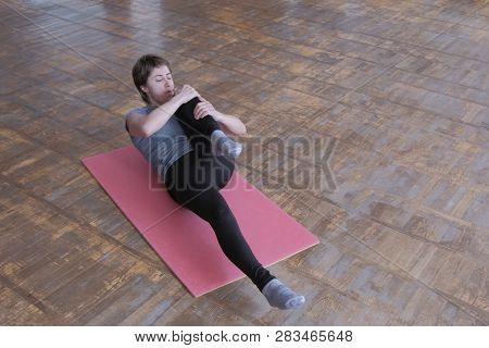 A Woman In A Gray T-shirt On A Pink Rug Raises Her Leg To Her Chest. Fitness Exercise To Strengthen 