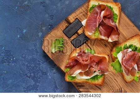 Bruschetta With Prosciutto, Salad Frieze And Cheese On The Kitchen Table. Snack. View From Above.