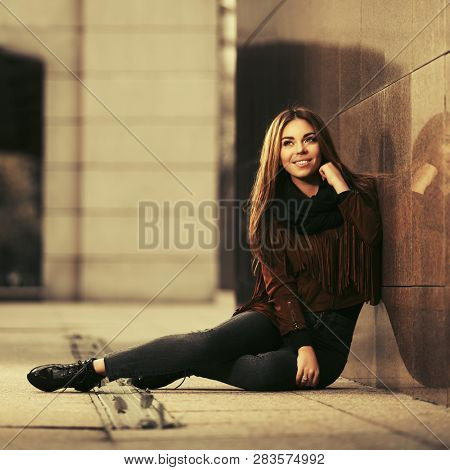 Happy young fashion woman sitting on city street sidewalk  Stylish female model wearing brown leather fringe suede jacket and dark blue jeans