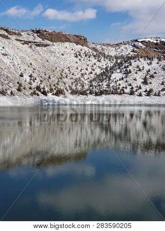 Snow Covered Hills, Trees, Clouds, And A Bright Blue Sky Reflect In The Partially Iced Over Prinevil
