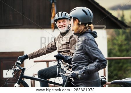Active Senior Couple With Electrobikes Standing Outdoors In Front Of A House.