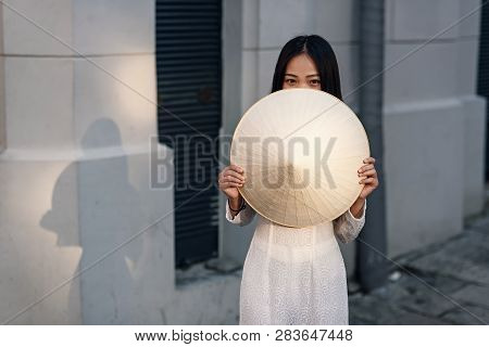 Cute Happy Asian Girl Dressed In Ao Dai Dress Closes Half Of Her Face With Vietnamese Conical Hat.