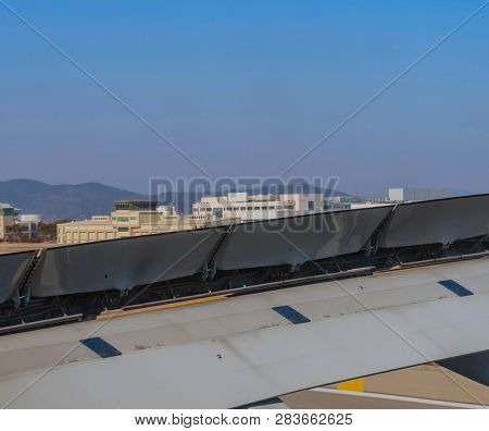 Aircraft Wing With Breaking Flaps Extended During Landing With Airport Buildings And Blue Sky In Bac