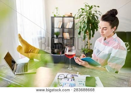 Side view portrait of relaxed young woman writing in journal with feet on desk, copy space