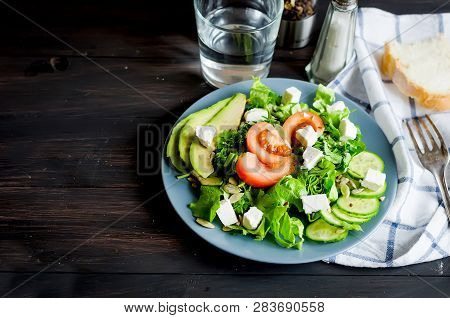 Fresh  Green Salad With Cucumber, Lettuce, Avocado And Tomatoes And Glass Water On A Dark Wooden Tab