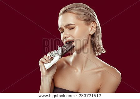 Blonde-haired Model Biting Chocolate Bar Standing Near Red Wall