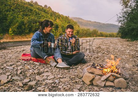 Two Young Couple Sitting Near Campfire Drinking Warm Up Tea And Talking. Lifestyle Concept