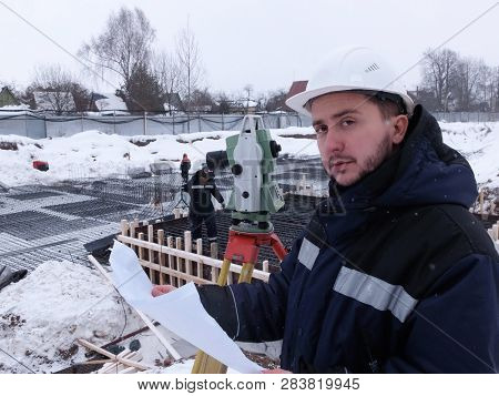 Bearded Geodetic Engineer-surveyor In White Hardhat Looks At The Camera Holding A Drawing Sheet Agai