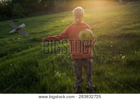 Boy Threw Away Paper Plane. Child Is Holding Yellow Ball. Walking At Sunset Before Bedtime. Happy Ch