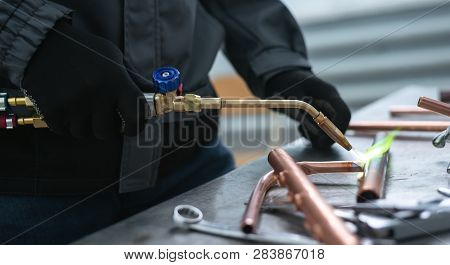 Worker Is Soldering A Pipe By A Blow Lamp On A Factory Workbench Background. Pipework.