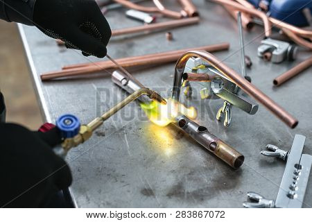 Worker Is Soldering A Pipe By A Blow Lamp On A Factory Workbench Background. Pipework.