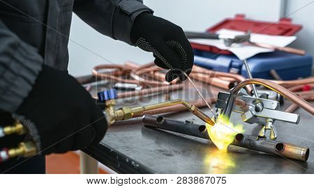 Worker Is Soldering A Pipe By A Blow Lamp On A Factory Workbench Background. Pipework.