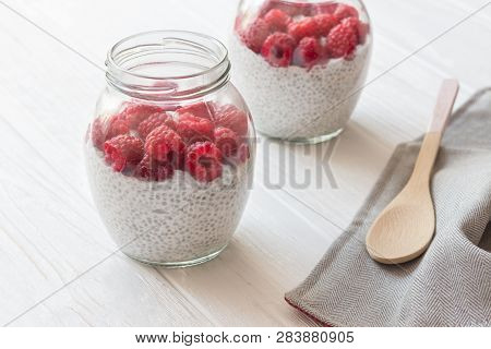 Glass Jars Of Pudding From Chia Seeds With Coconut Milk And Raspberry Berries In White Wooden Table.