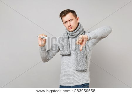 Upset Young Man In Gray Sweater, Scarf Holding Thermometer, Showing Thumb Down Isolated On Grey Wall