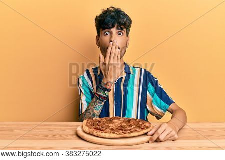 Young hispanic man eating italian pizza sitting on the table covering mouth with hand, shocked and afraid for mistake. surprised expression 