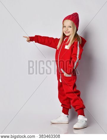 Cute Smiling Blonde Kid Girl In Red Overall Jumpsuit And Winter Knitted Hat Stands With Her Hand In 