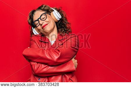 Middle age beautiful woman listening to music using headphones over red background hugging oneself happy and positive, smiling confident. Self love and self care