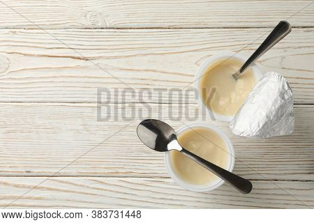 Plastic Cups Of Yogurt And Spoons On White Wooden Background