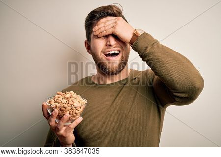 Young blond man with beard and blue eyes holding bowl with healthy salty peanuts stressed with hand on head, shocked with shame and surprise face, angry and frustrated. Fear and upset for mistake.