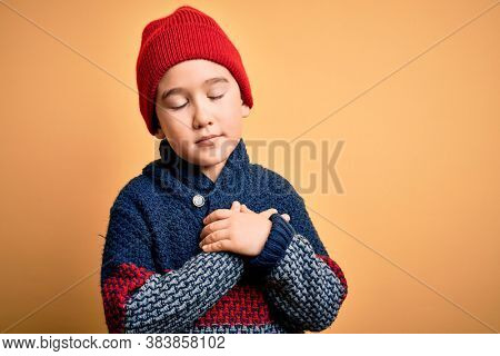 Young little boy kid wearing wool cap and winter sweater over yellow isolated background smiling with hands on chest with closed eyes and grateful gesture on face. Health concept.