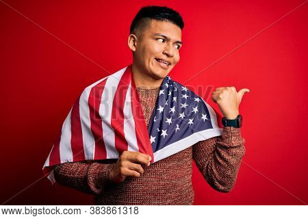 Young handsome latin patriotic man wearing united states of america falg pointing and showing with thumb up to the side with happy face smiling