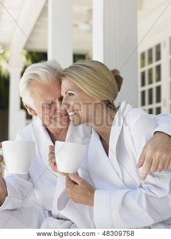 Romantisk midterste ældre par i badekåber med kaffekopper sidder på verandaen