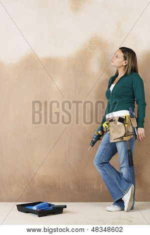 มุมมองด้านข้างแบบยาวของสาวที่มี toolbelt และสว่านพิงกับผนัง