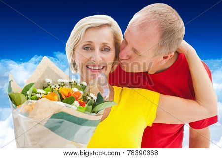 Mature man kissing his partner holding flowers against bright blue sky over clouds