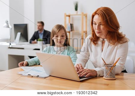 Interested in details. Curious bright productive girl watching her mom working on a report and handing her the diagrams she needing