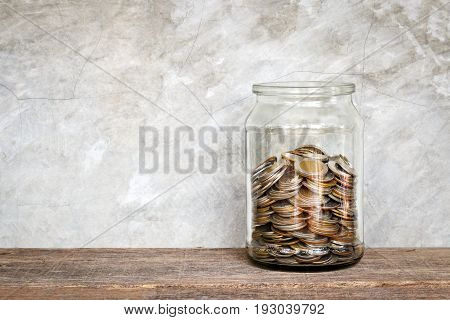 Coins in a glass jar on old wooden floor with grunge wall saving money concept.