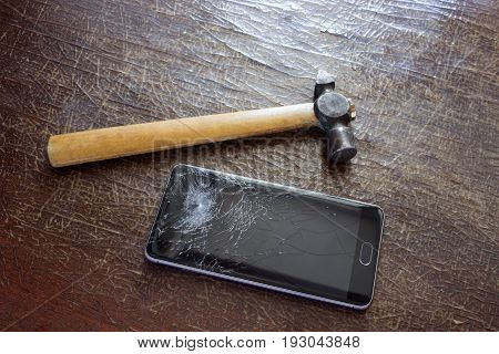 A hammer on a cracked table next to the phone with a broken screen