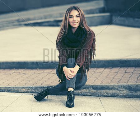 Happy young fashion woman sitting on city sidewalk. Stylish female model in leather fringe suede jacket and blue jeans