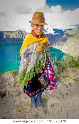 QUITO, ECUADOR - NOVEMBER, 25 2016: Unidentified indigenous old woman wearing a typical andean clothes in front of the Quilotoa lake with a beautiful magenta color of the Quilotoa caldera. Quilotoa is the western volcano in Andes range and is located in a