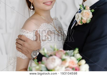 Handsome groom in dark grey suit gently holding beautiful smiling bride with white veil and bridal bouquet of roses and peonies by the shoulders and kissing her at the wedding ceremony.