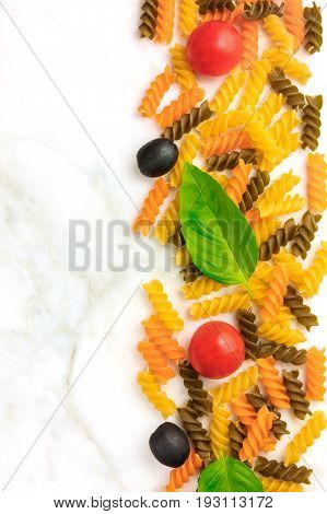 Pasta salad ingredients on white marble with a place for text. Rotini, cherry tomatoes, fresh basil leaves, and black olives