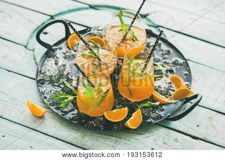 Refreshing cold alcoholic summer citrus cocktail with orange, peppermint and crushed ice in stemless glasses on dark tray over light wooden background, selective focus, horizontal composition