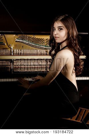 beautiful young woman in a black dress with an open back standing nere the piano on a dark background.