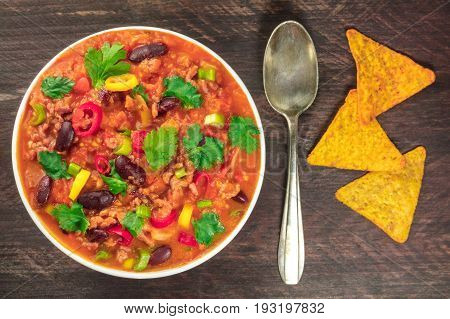 A photo of chilli con carne, a Mexican dish with red beans, coriander leaves, ground meat, and chilli peppers, on a dark rustic texture with nachos and a spoon, shot from above