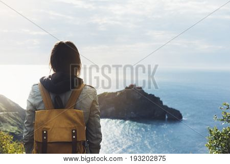 Hipster young girl with backpack enjoying sunset on seascape on peak mountain. Tourist traveler on background valley landscape view mockup. Hiker looking sunlight ocean in trip holiday in basque island Gaztelugatxe