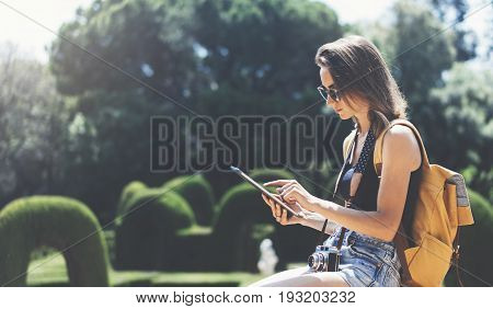 Hipster young girl with bright backpack and vintage camera using tablet or holding gadget planning travel plan. View tourist traveler with sunglasses on background Parc Laberint Barcelona. Mockup for text message