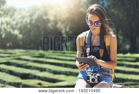 Hipster young girl with bright backpack and vintage camera using tablet or holding gadget planning travel plan. View tourist traveler on background Parc Laberint Barcelona. Mock up for text message