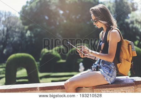Hipster young girl with bright backpack and vintage camera using tablet or holding gadget planning travel plan. View tourist traveler with on background Parc Laberint Barcelona. Mock up for text message