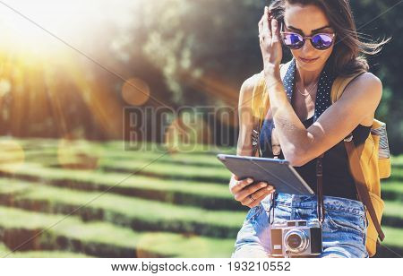 Hipster young girl with bright backpack and vintage camera using tablet or holding gadget planning travel plan. View tourist traveler on background Parc Laberint Barcelona. Mock up for text message