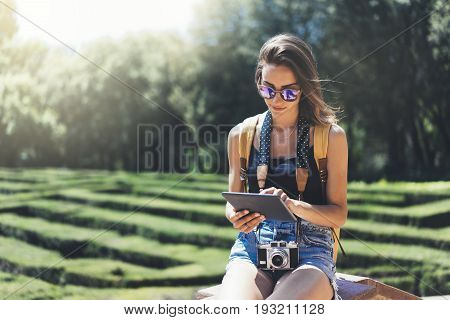 Hipster young girl with bright backpack and vintage camera using tablet or holding gadget planning travel plan. View tourist traveler on background Parc Laberint Barcelona. Mock up for text message