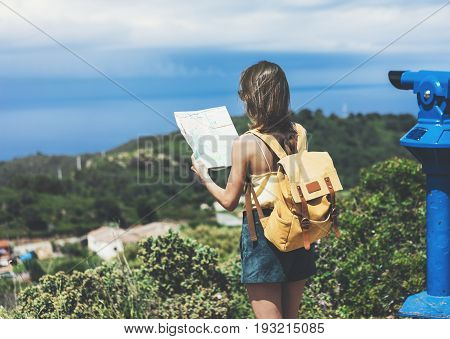 Hipster young girl with bright backpack looking at a map and poining hand the travel plan. View from the back of the tourist traveler on background mountain sea coin operated binoculars. Mock up for text message