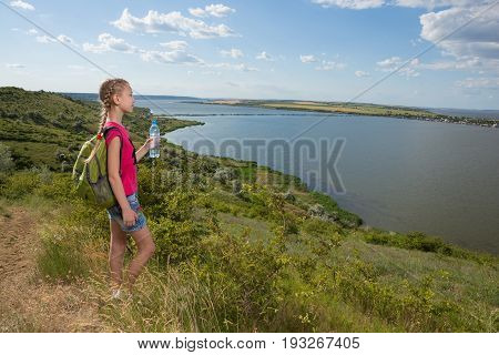 Girl with a backpack standing on the background of the lake and drinking water, sunny day